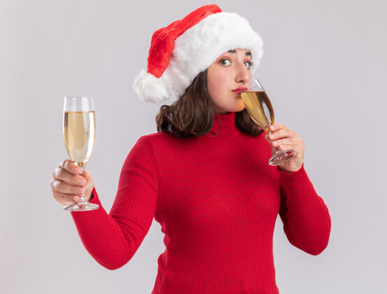 年轻穿着红色毛衣 戴着圣诞帽的年轻女孩 手拿两杯香槟 站在白色的背景下快乐而积极地喝着帽子眼镜积极的