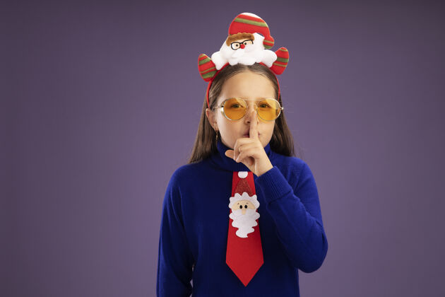 制作小女孩穿着蓝色高领毛衣 打着红色领带 头上戴着有趣的圣诞戒指 看着摄像机 手指放在嘴唇上 站在紫色背景上做着沉默的手势领带圣诞节搞笑