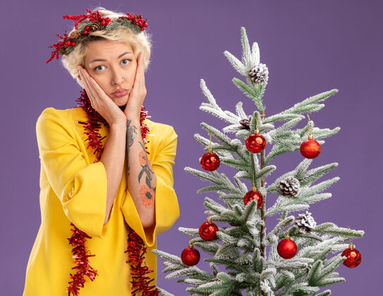 花环无聊的金发女郎头戴圣诞花环 脖子上戴着金属丝花环 站在装饰好的圣诞树旁 手放在脸上 孤立地站在紫色的墙上花环头女人