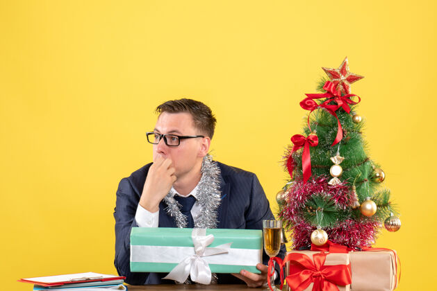 办公室前视图：体贴的男人坐在圣诞树旁的桌子旁 黄色的礼物坐着电脑树