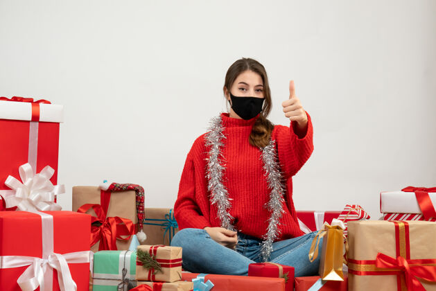 肖像穿着红色毛衣的年轻女孩做着竖起大拇指的招牌围坐在礼物旁边 白色的面具上戴着黑色的面具服装圣诞节礼物