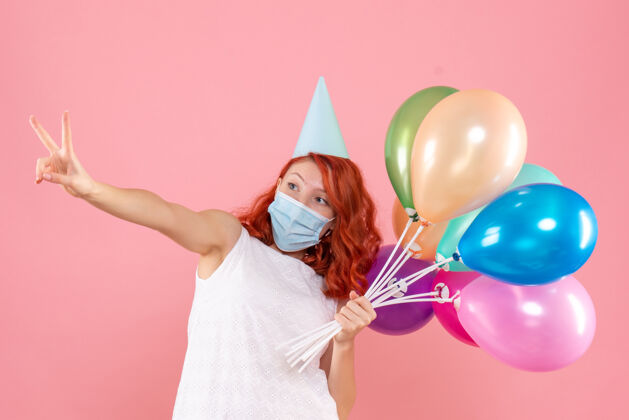 欢乐前视图中的年轻女子拿着五颜六色的气球在无菌面具上粉红色的墙壁举行面具前面
