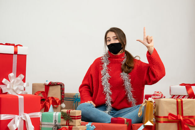 面具穿着红色毛衣 戴着黑色面具的年轻女孩正在做手指枪的手势 围坐在白色的礼物上坐着毛衣周围