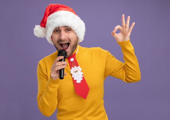 相机自信的年轻白种人戴着圣诞帽打着领带拿着麦克风唱歌看着镜头做ok标志孤立在紫色背景上自信帽子看着