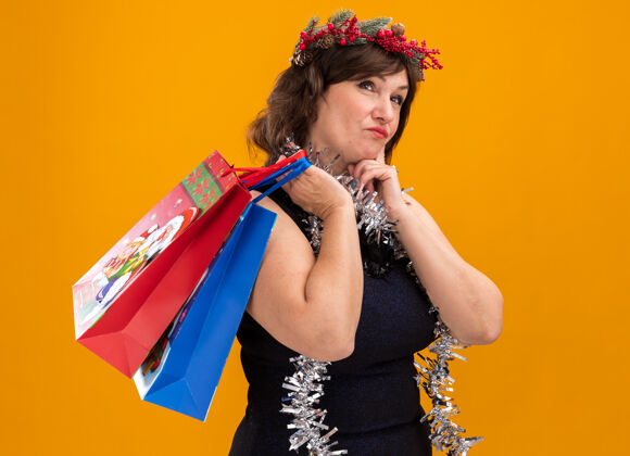 脖子体贴的中年妇女头戴圣诞花环 脖子上戴着金属丝花环 手持圣诞礼品袋站在侧视图上下巴圣诞包