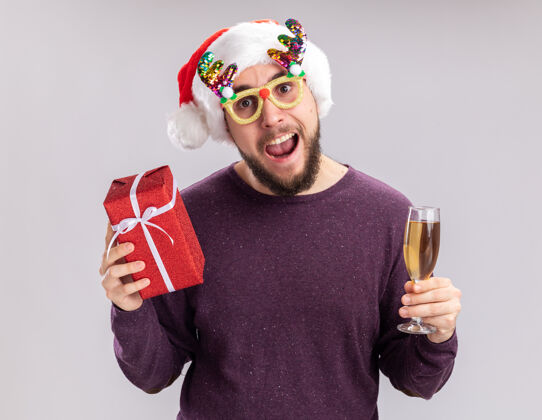 帽子快乐的年轻人穿着紫色毛衣 戴着圣诞帽 戴着滑稽的眼镜 手里拿着一杯香槟和礼物年圣诞毛衣