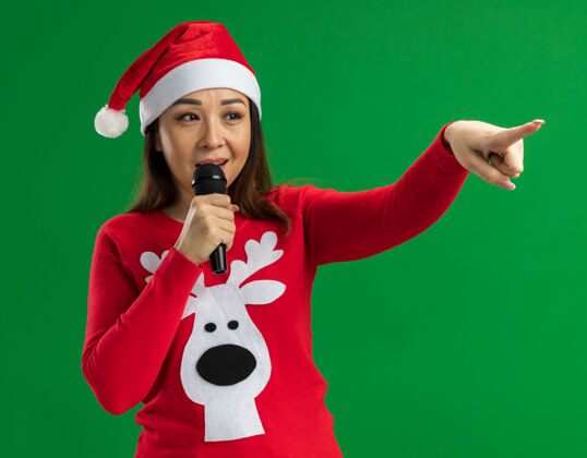 脸戴着圣诞老人帽 穿着红色毛衣的年轻女子对着麦克风说话 一边看着一边开心的脸 一边站在绿色背景上指指点点麦克风圣诞老人帽子