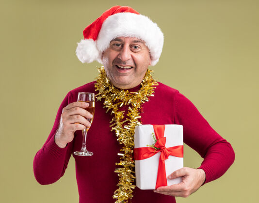 圣诞老人快乐的中年男人戴着圣诞老人帽 脖子上戴着金属丝 手里拿着圣诞礼物 手里拿着一杯香槟 站在绿色的背景下 面带微笑地看着相机周围杯子礼物