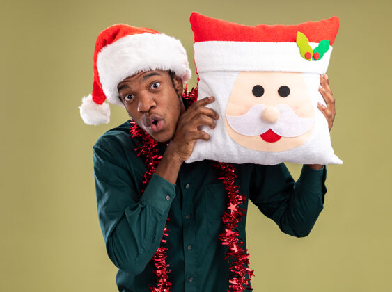 花环一个非洲裔美国人戴着圣诞帽 戴着花环 抱着圣诞枕头 站在绿色的背景下惊讶地看着相机帽子圣诞节惊讶