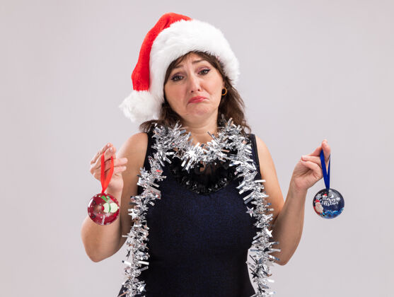 圣诞节困惑的中年妇女戴着圣诞帽 脖子上戴着金属丝花环 手里拿着圣诞饰品 看着隔离在白色背景上的相机中年帽子圣诞老人
