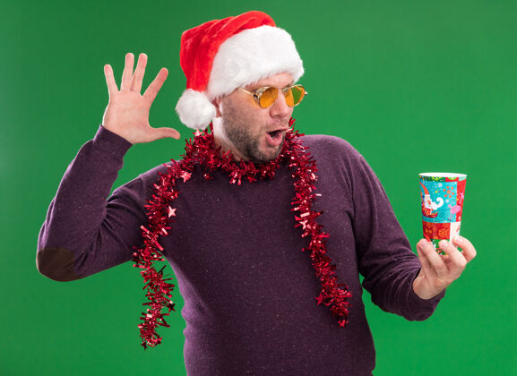 圣诞老人令人印象深刻的中年男子戴着圣诞帽 脖子上戴着金箔花环 戴着眼镜 举着塑料圣诞杯 看着绿色的墙上孤立的手男人花环圣诞节