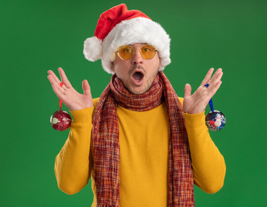 高领毛衣一个穿着黄色高领毛衣 戴着圣诞帽 戴着眼镜 手里拿着圣诞树玩具的年轻人站在绿色的墙上 惊讶不已站着帽子惊讶
