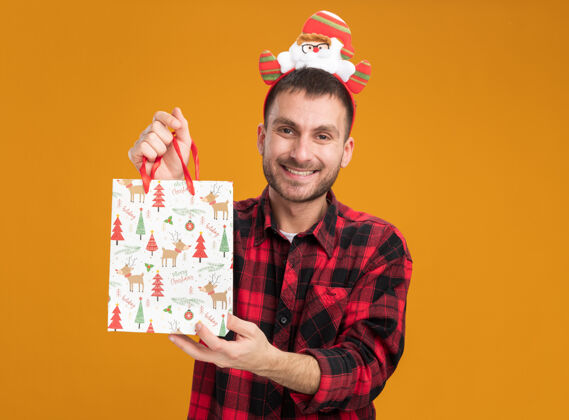 戴着快乐的白人年轻人戴着圣诞老人的头带 拿着圣诞礼品袋 隔离在橙色的墙上 留着复制空间圣诞节年轻人男人