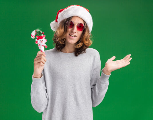 圣诞节年轻人戴着圣诞老人帽 戴着红色眼镜 手持圣诞糖果手杖 看着镜头 微笑着 手臂站在绿色的背景上 欢快地呈现圣诞老人糖果眼镜