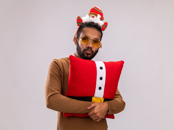 圣诞老人一个非洲裔美国人 穿着棕色毛衣 头戴圣诞老人圈 打着有趣的红色领带 抱着圣诞枕头 站在白色背景下困惑地看着摄像机非洲枕头抱着