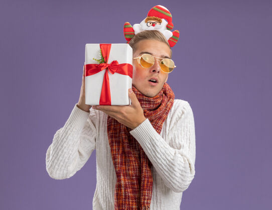 帅气好奇的年轻帅哥戴着圣诞老人的头带和围巾拿着礼包靠近头部孤立的紫色墙壁上有复制空间近圣诞老人包