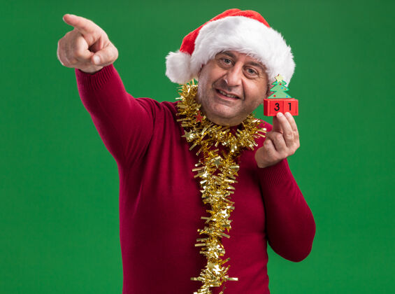 年龄快乐的中年男人戴着圣诞老人帽子 脖子上戴着金属丝 手里拿着新年礼物的玩具立方体 微笑着用食指着站在绿色背景上的一边立方体日期围着