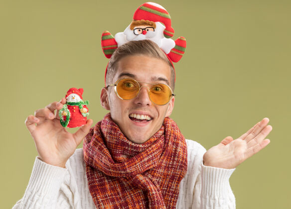 年轻印象深刻的年轻帅哥戴着圣诞老人的头带和围巾拿着雪人的圣诞饰品显示空手孤立在橄榄绿的墙上圣诞老人展示围巾