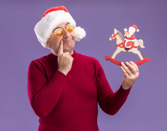 抱着中年男子戴着圣诞老人帽 戴着黄色眼镜 手里拿着圣诞玩具 站在紫色的背景上困惑地抬头看圣诞节困惑中间