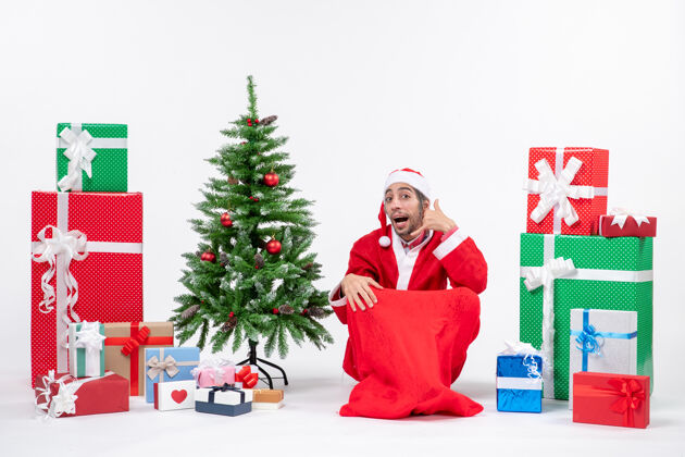 圣诞老人年轻人打扮成圣诞老人 拿着礼物和装饰好的圣诞树坐在地上做着叫我的手势圣诞老人礼物庆祝