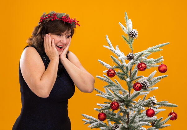 轮廓令人印象深刻的中年妇女头戴圣诞花环 脖子上戴着金属丝花环 站在装饰过的圣诞树旁的侧视图上站着花圈快乐