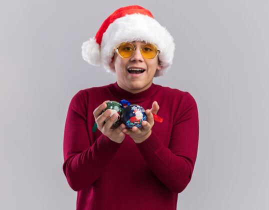 帽子快乐的年轻人戴着圣诞帽 戴着眼镜 手里拿着圣诞树上的球 背景是白色的舞会圣诞树快乐