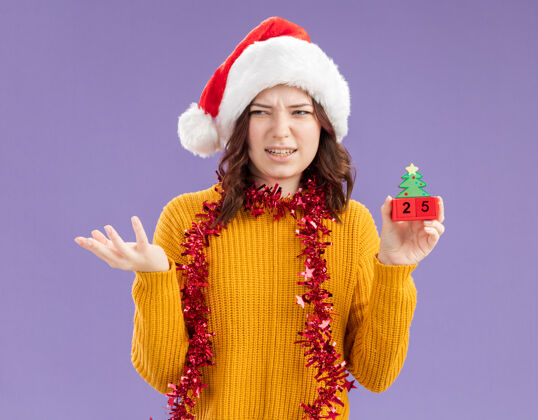 装饰品恼怒的年轻斯拉夫女孩戴着圣诞帽 脖子上戴着花环 手里拿着圣诞树饰物 看着紫色背景上孤立的一面 还有复制空间帽子斯拉夫周围