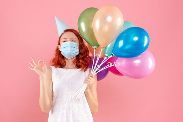 快乐正面图是粉红色墙上戴着消毒面具手持彩色气球的年轻女子粉色年轻气球