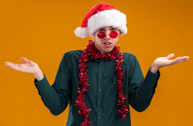 手笨手笨脚的年轻金发男子戴着圣诞帽 戴着眼镜 脖子上戴着金属丝花环 看着摄像机 两手空空的 孤立在橙色背景上脖子空年轻