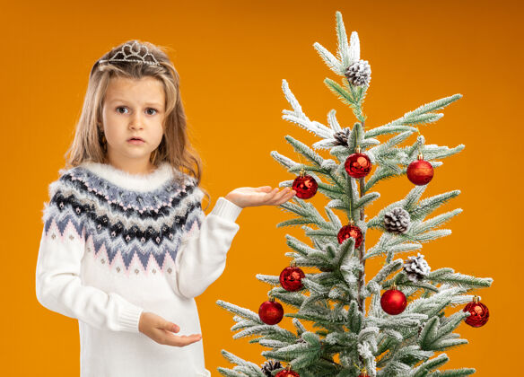 小站在圣诞树旁的小女孩戴着头饰 脖子上有花环 侧面孤立在橙色背景上点圣诞节树