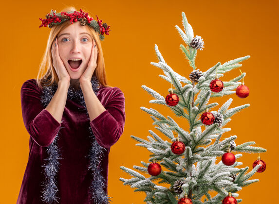 脸颊站在圣诞树旁的年轻漂亮的女孩穿着红色的裙子 脖子上戴着花环 手放在脸颊上 橙色的背景隔离开来年轻女孩脖子