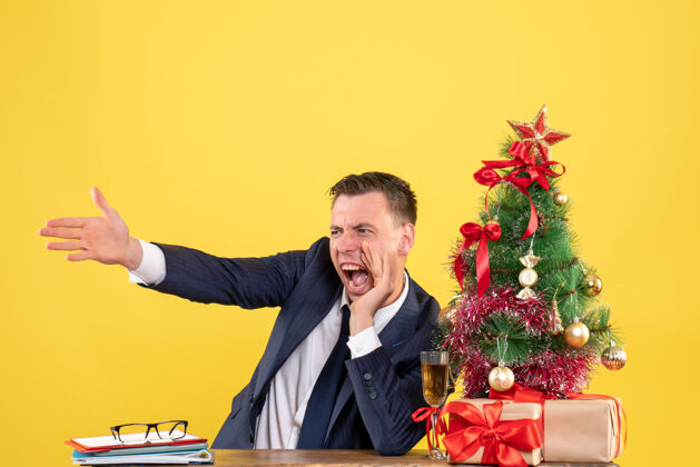 商人愤怒的男人坐在圣诞树旁大声喊叫 黄色墙上挂着礼物男人喊叫圣诞节