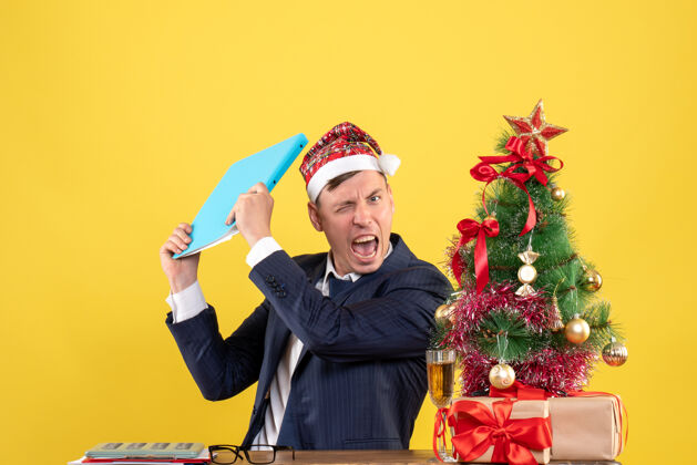 前面前视图愤怒的商人拿着文件文件坐在圣诞树旁的桌子旁 黄色的墙上摆着礼物男人文件