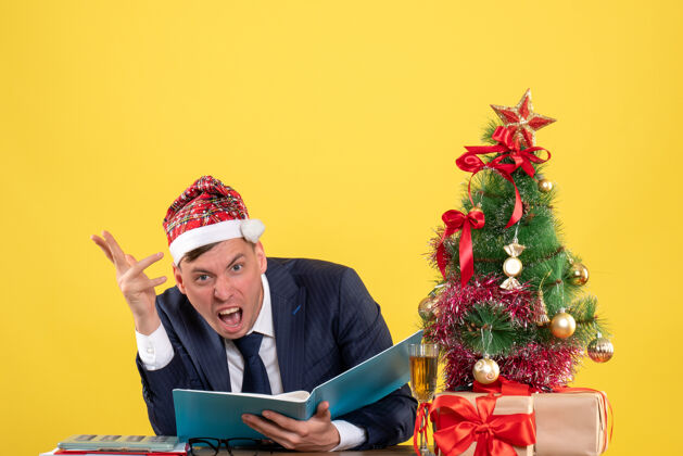 圣诞老人愤怒的商人戴着圣诞帽坐在圣诞树旁的桌子前 黄色墙上挂着礼物人桌子商人