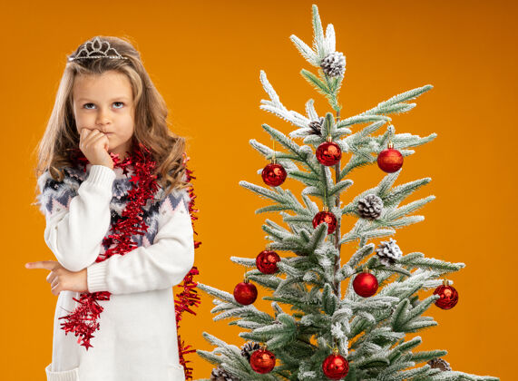 看想着抬头看看站在圣诞树旁戴着头饰脖子上戴着花环的小女孩 手放在下巴上 孤立在橙色背景上花环圣诞节脖子