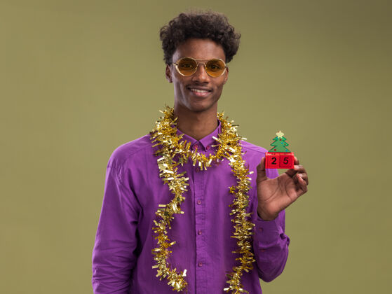 脖子微笑的非洲裔美国年轻人戴着眼镜 脖子上戴着金属丝花环 手里拿着圣诞树玩具和日期 看着橄榄绿背景上孤立的相机非裔美国人微笑树