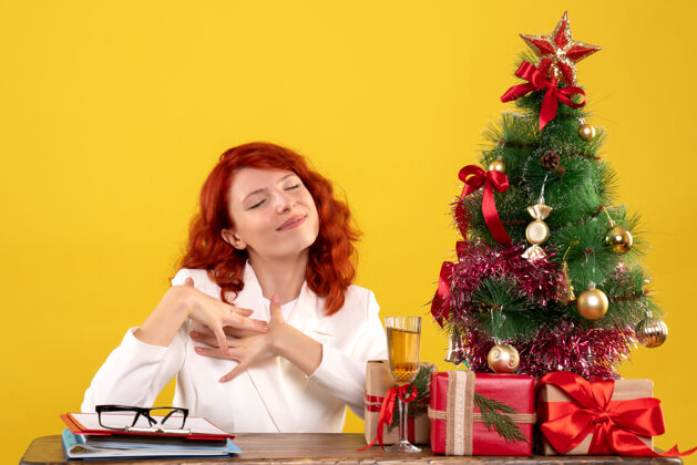 情感女工人坐在桌子后面 手里拿着圣诞礼物和黄色的圣诞树桌子安排坐着