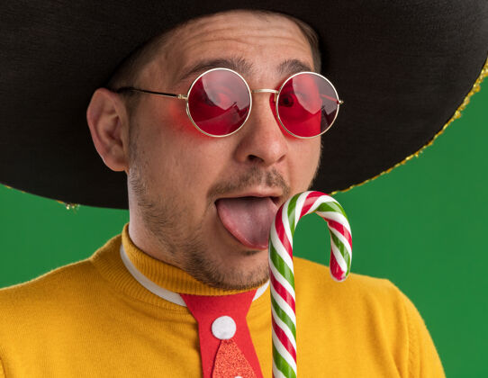 站着一个穿着黄色高领毛衣 戴着眼镜 戴着滑稽的红色领带 戴着黑色帽子 拿着糖果手杖的年轻人看着镜头 在绿色的背景下 他露出一张幸福的脸 伸出舌头脸搞笑糖果