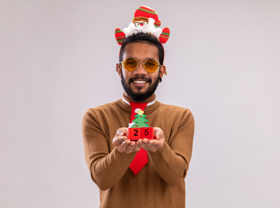 日期快乐的非洲裔美国人 身穿棕色毛衣 头戴圣诞老人戒指 打着有趣的红色领带 在白色背景上 他站着一个快乐的微笑 展示着玩具方块和日期25立方体头非洲