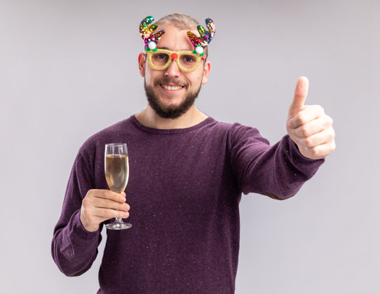 香槟一个穿着紫色毛衣戴着滑稽眼镜的快乐的年轻人 手里拿着一杯香槟 面带微笑地看着相机 脚上穿鞋 竖起大拇指站在白色的背景上穿鞋拿着搞笑