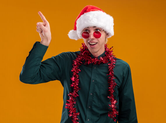 花环令人印象深刻的年轻金发男子戴着圣诞帽 戴着眼镜 脖子上戴着金箔花环 看着一边指着橙色背景上的孤立年轻手指印象深刻