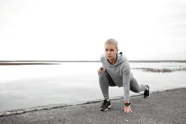 健康穿着时髦运动服的漂亮的自我克制的高加索女跑步者在河岸上摆出孤立的姿势 伸展双腿 热身肌肉 然后一大早在户外跑步锻炼运动员湖泊健身