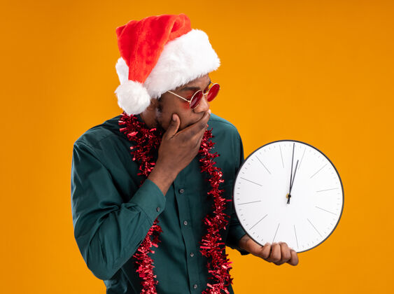 美国人非洲裔美国人戴着圣诞帽 戴着花环 戴着太阳镜 拿着钟 站在橙色的背景上惊讶地看着它帽子站着钟表