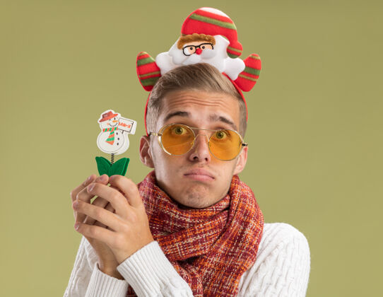 帅哥悲伤的年轻帅哥戴着圣诞老人的头带和围巾看着相机拿着橄榄绿背景上孤立的雪人玩具围巾小伙子雪人