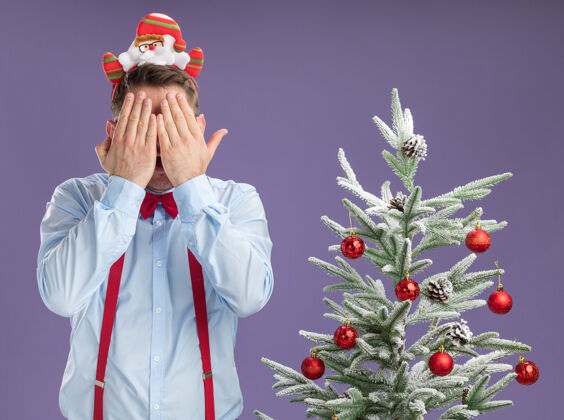 站着戴着吊带领结的年轻人站在圣诞树旁 戴着圣诞老人和红眼镜 手放在紫色背景上遮住脸轮辋弓领带