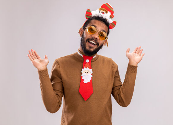毛衣一个身穿棕色毛衣 头戴圣诞老人戒指 打着有趣的红色领带的非裔美国人站在白色背景下 看着摄像机 开心地微笑着搞笑快乐站立
