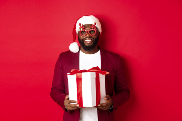 正式圣诞节英俊的非洲裔美国人戴着派对眼镜 戴着圣诞帽 手里拿着新年礼物单身模特促销
