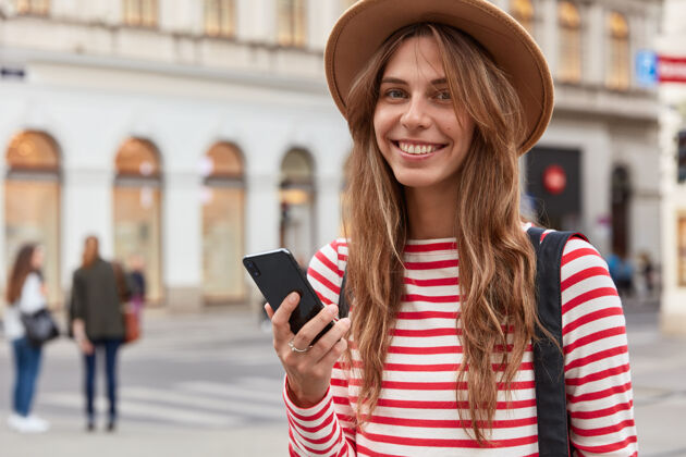 信息快乐的女游客使用旅游博客上的信息 手持智能手机 在城市街道上行走 戴着时髦的帽子和条纹套头衫姿势城镇街道