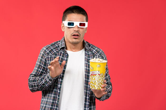 男性正面图：戴着d型太阳镜的年轻男子拿着爆米花站在浅红色的墙上看男性电影肖像电影院观点