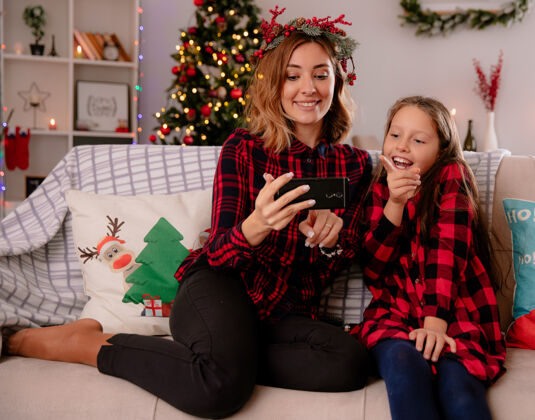 坐高兴的妈妈和女儿坐在沙发上看电话 在家里享受圣诞节的时光享受圣诞节沙发
